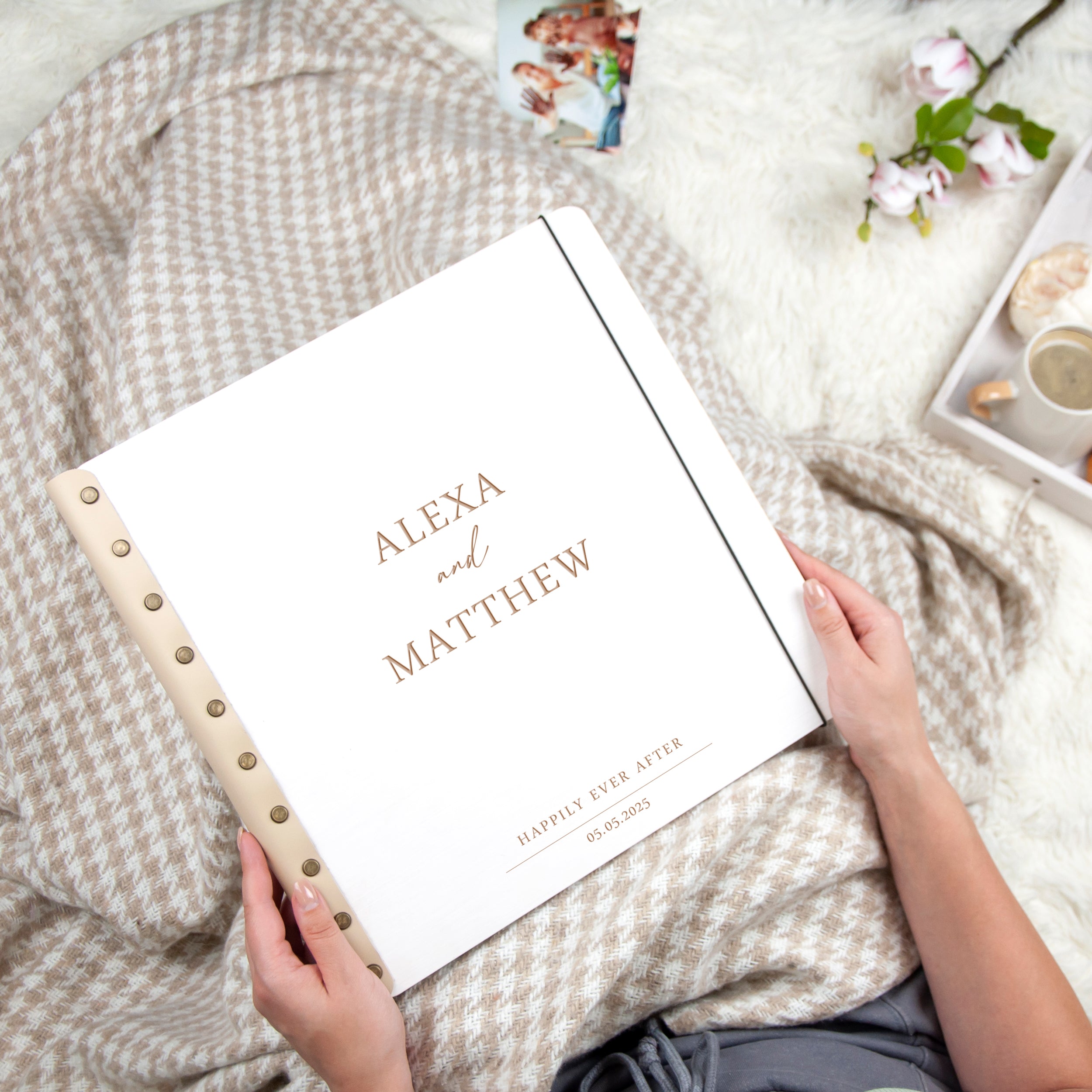 A person is holding a white book with the title "ALEXA and MATTHEW" printed on it, sitting on a blanket with a floral pattern.
