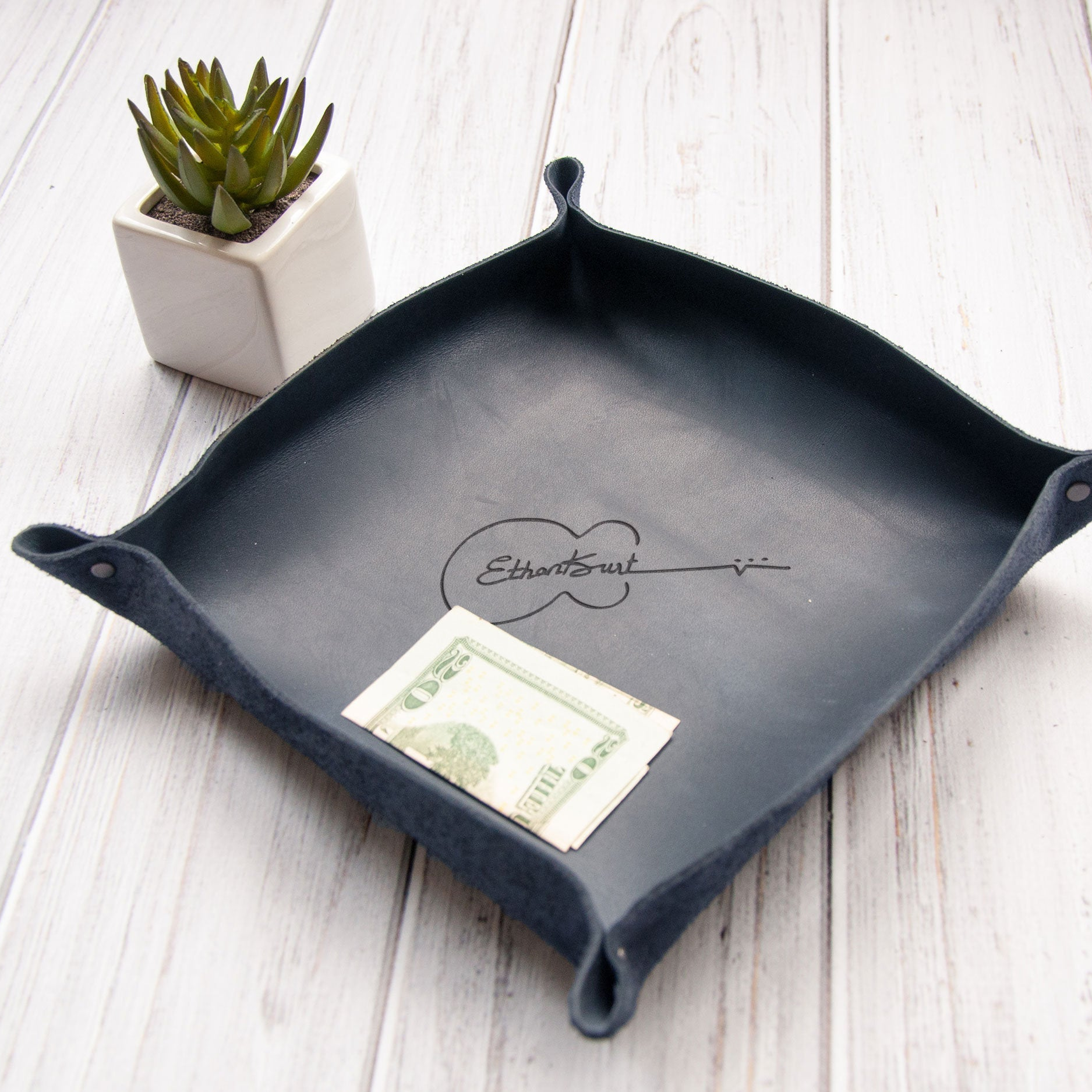 A black leather tray with a guitar-shaped design, placed on a white wooden surface next to a small potted succulent plant and a US dollar bill.