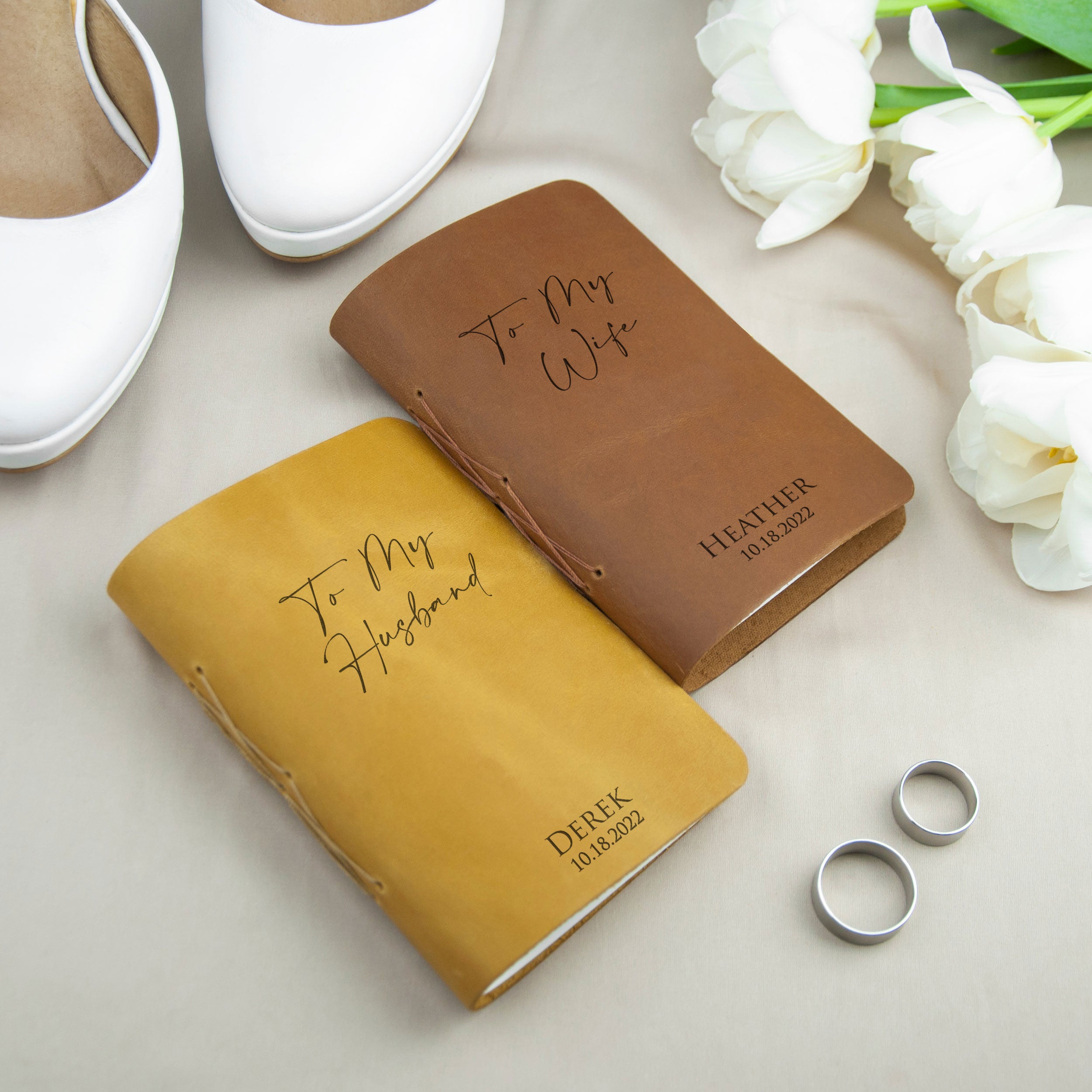 A pair of leather-bound notebooks with personalized messages, a pair of wedding rings, and a pair of white shoes are arranged on a table.
