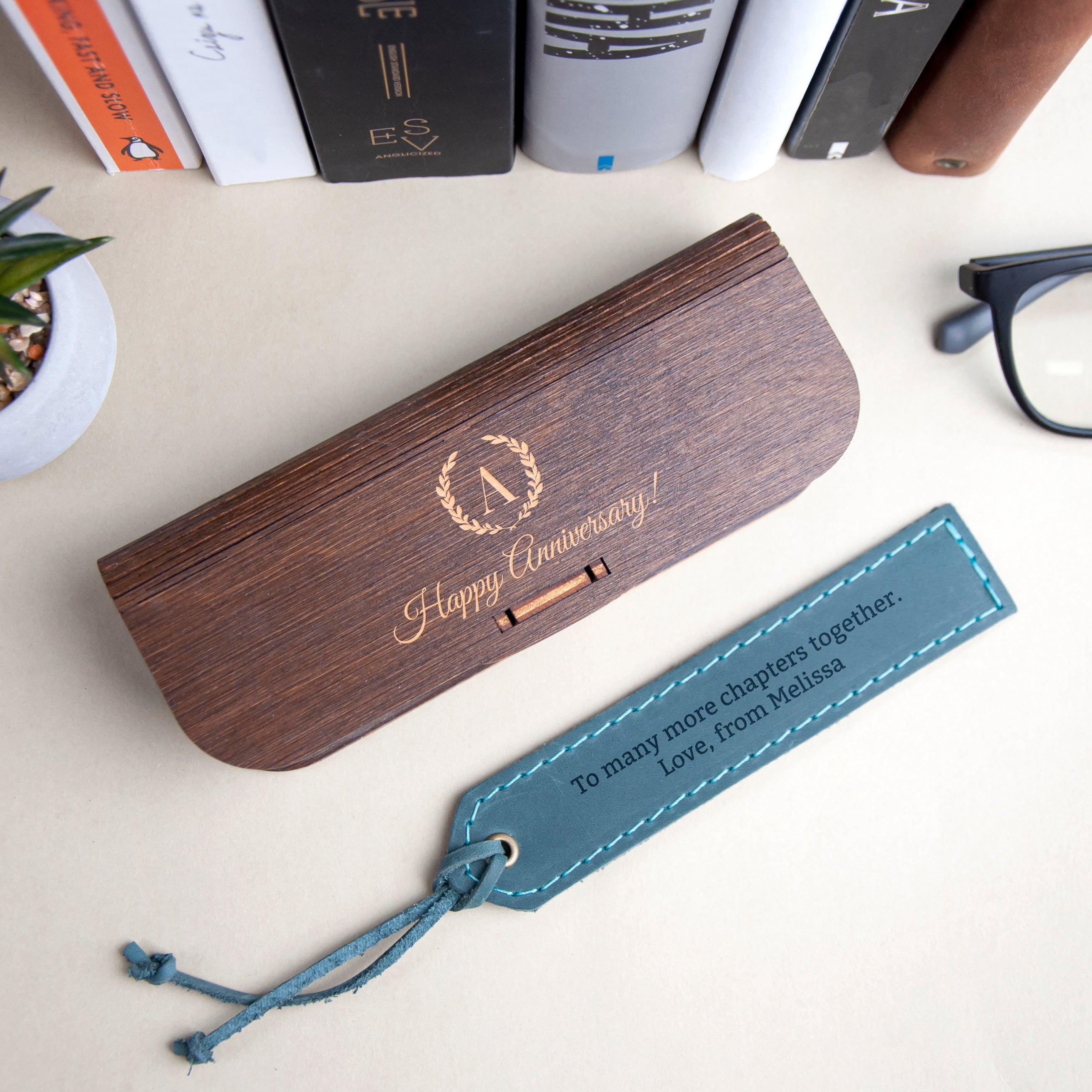 a wooden box with a gold embossed "Happy Anniversary!" and a blue tag with a message that reads "To many more chapters together. Love, from Melissa". The box and tag are placed on a desk, surrounded by books and a pair of glasses.