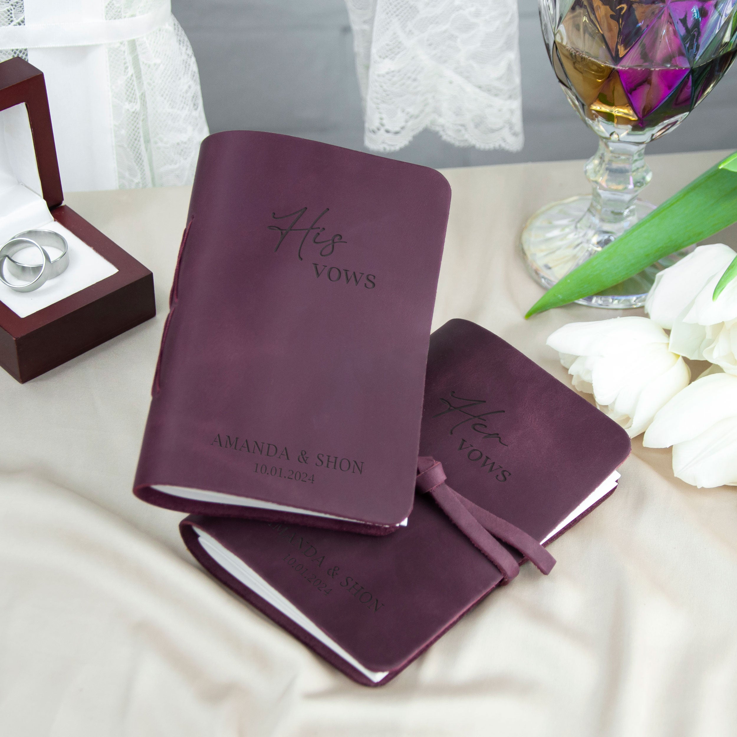 two purple leather-bound books with the text "His & hers" printed on them, placed on a table with a glass of wine, white flowers, and a ring box.