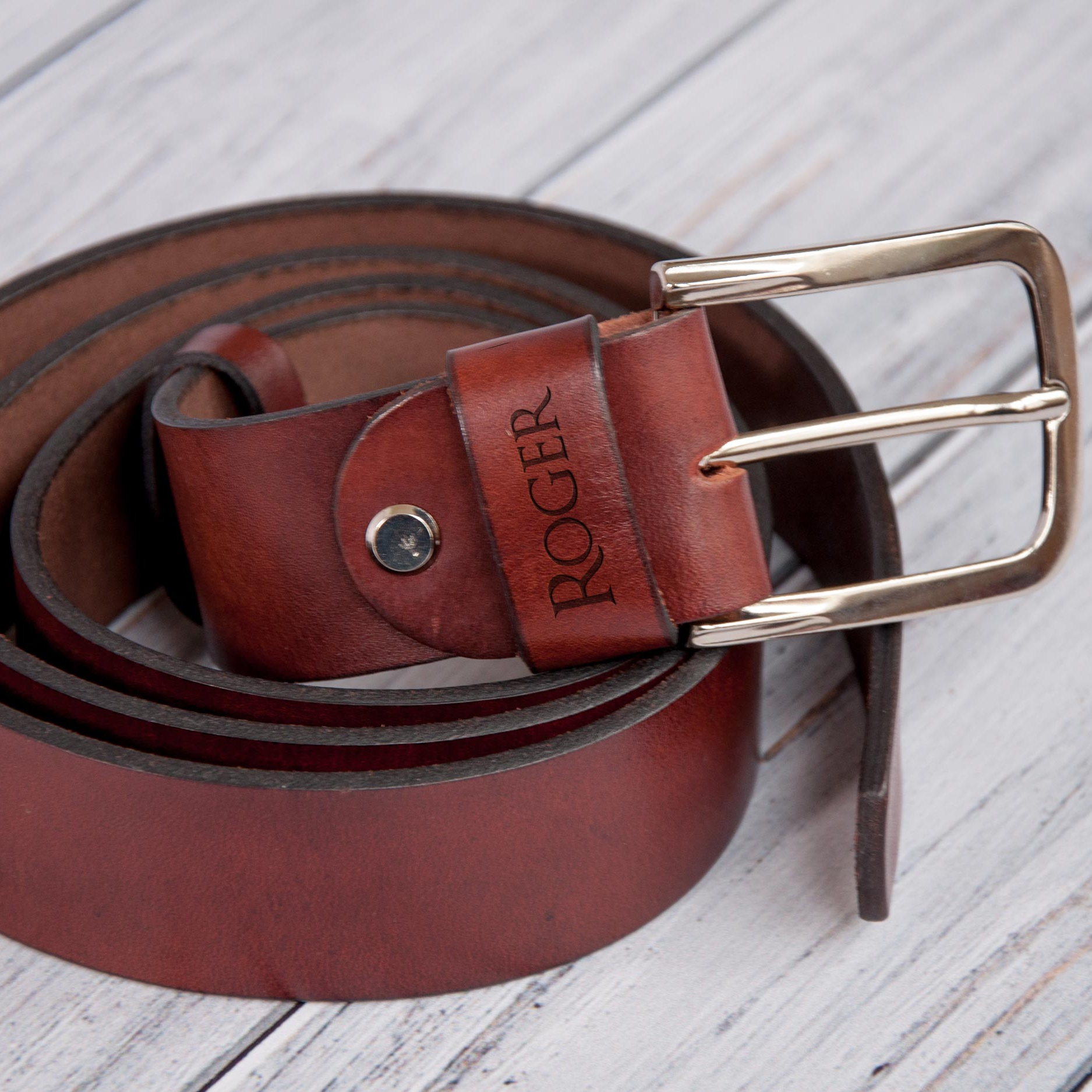 A brown leather belt with a personalized name engraved on it, resting on a wooden surface.