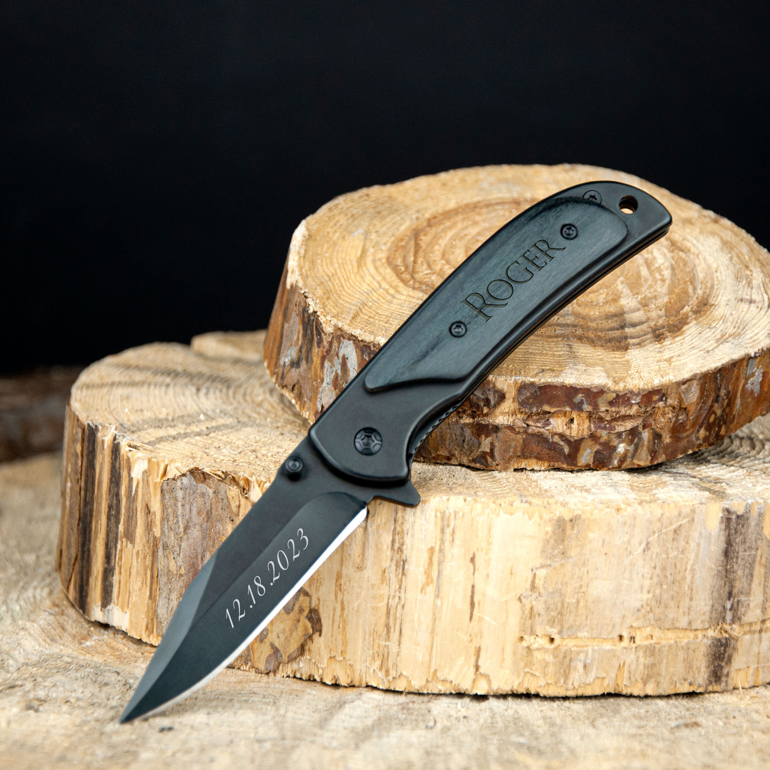 A black folding knife with the name "ROGER" engraved on it, resting on a wooden stump.