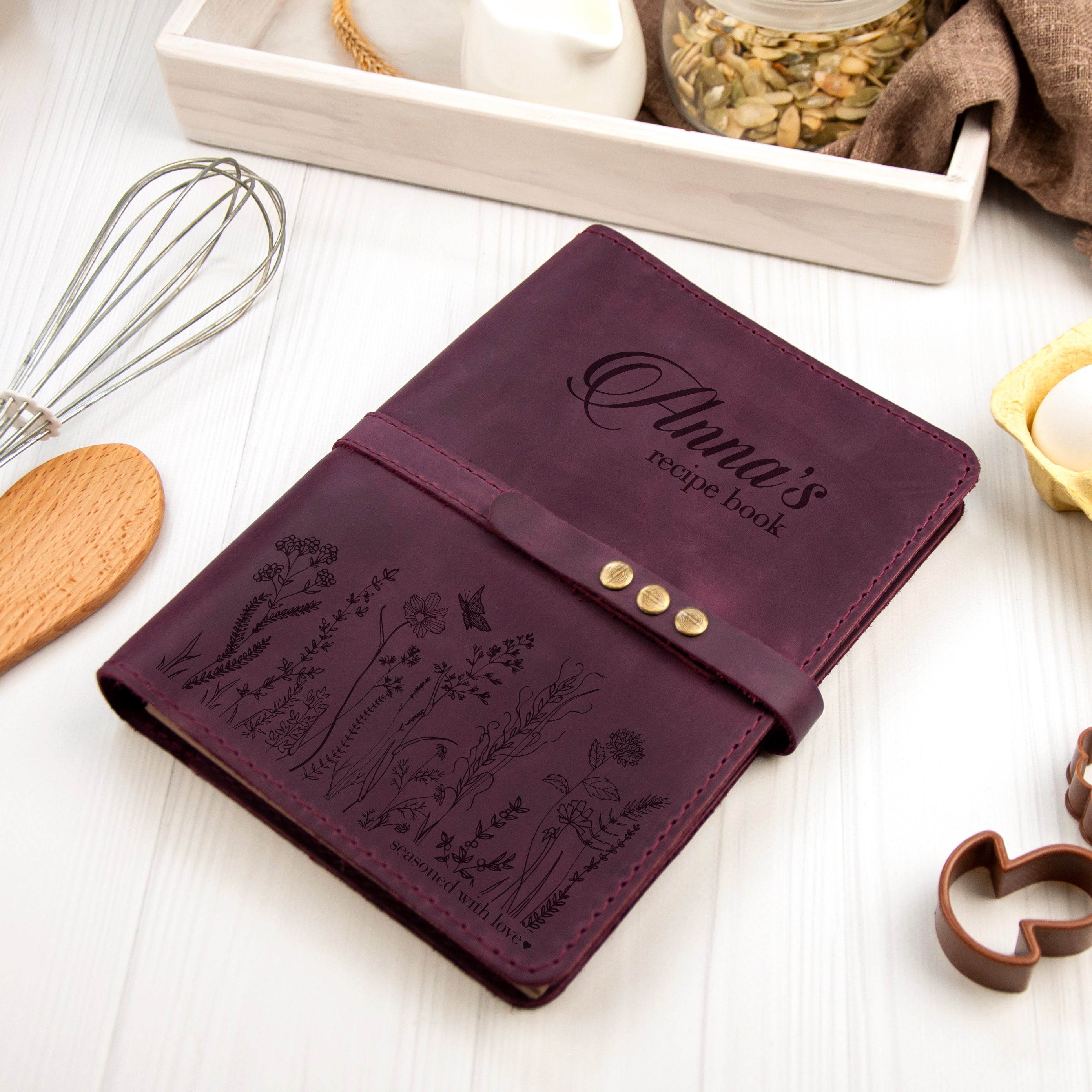 A purple leather recipe book with a metal clasp and a wooden spoon is placed on a white surface, surrounded by various kitchen items such as a whisk, a jar of seeds, and a cookie cutter.