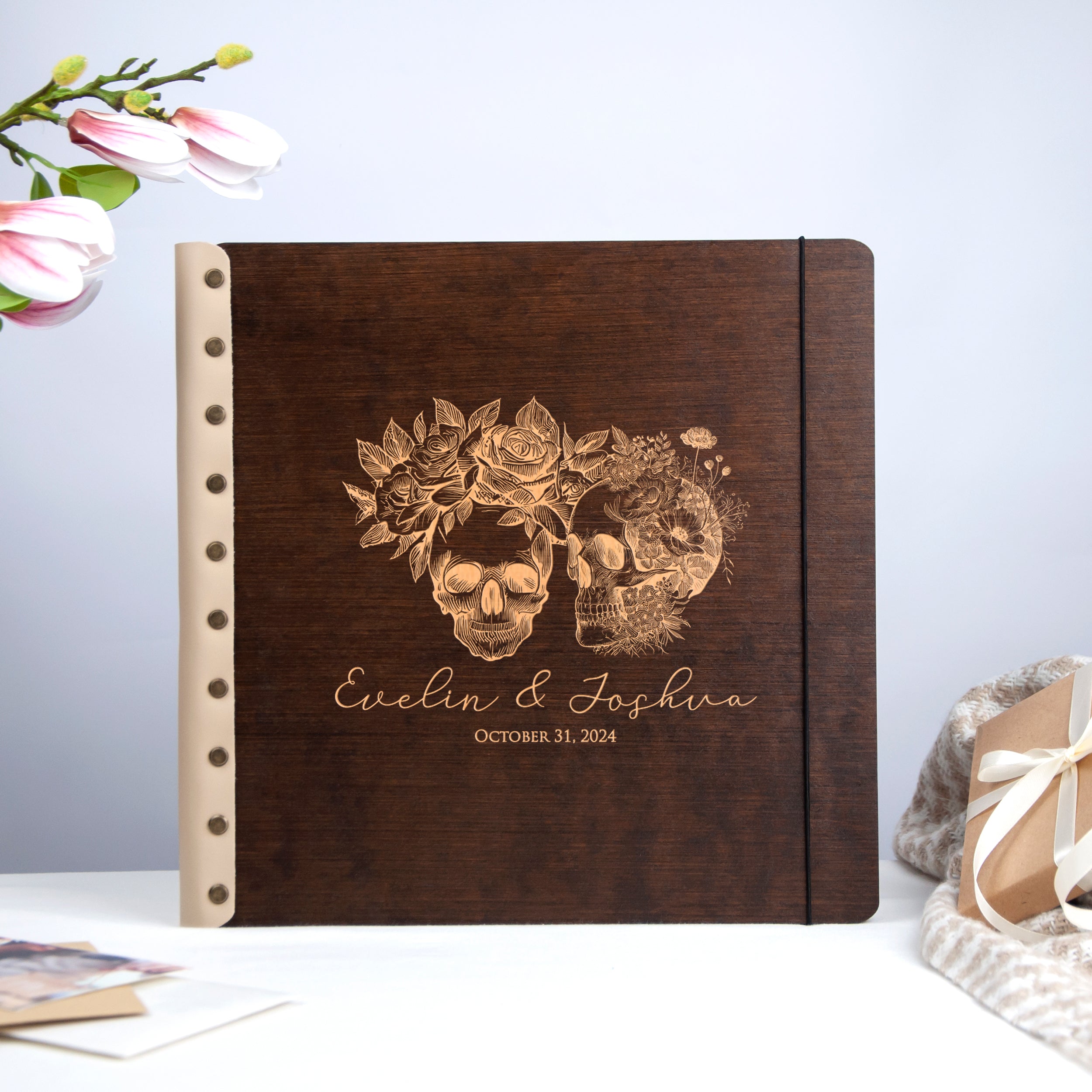 A wooden album with a floral design and the names 'Evelin & Joshua' printed on it, placed on a table with a gift bag and some flowers in the background.