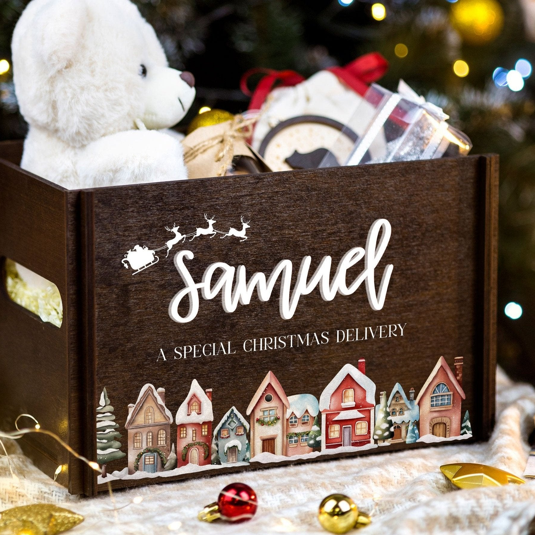 A wooden box with a teddy bear inside, decorated with a winter village scene and the name "Samuel" written on it.