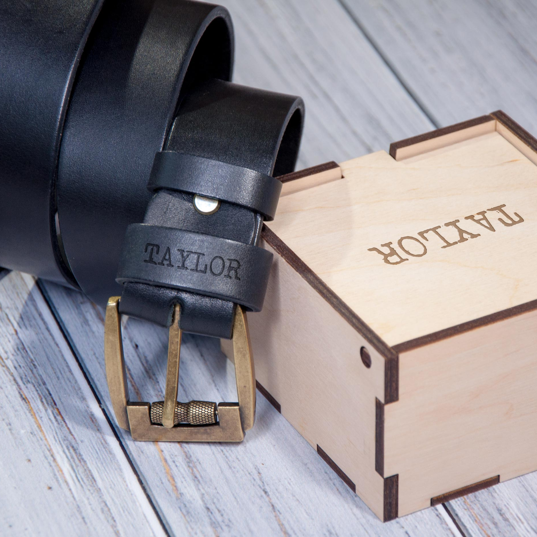 A black leather belt with the name "TAYLOR" engraved on it, along with a wooden box with the word "TAYLOR" engraved on it, both resting on a wooden surface.