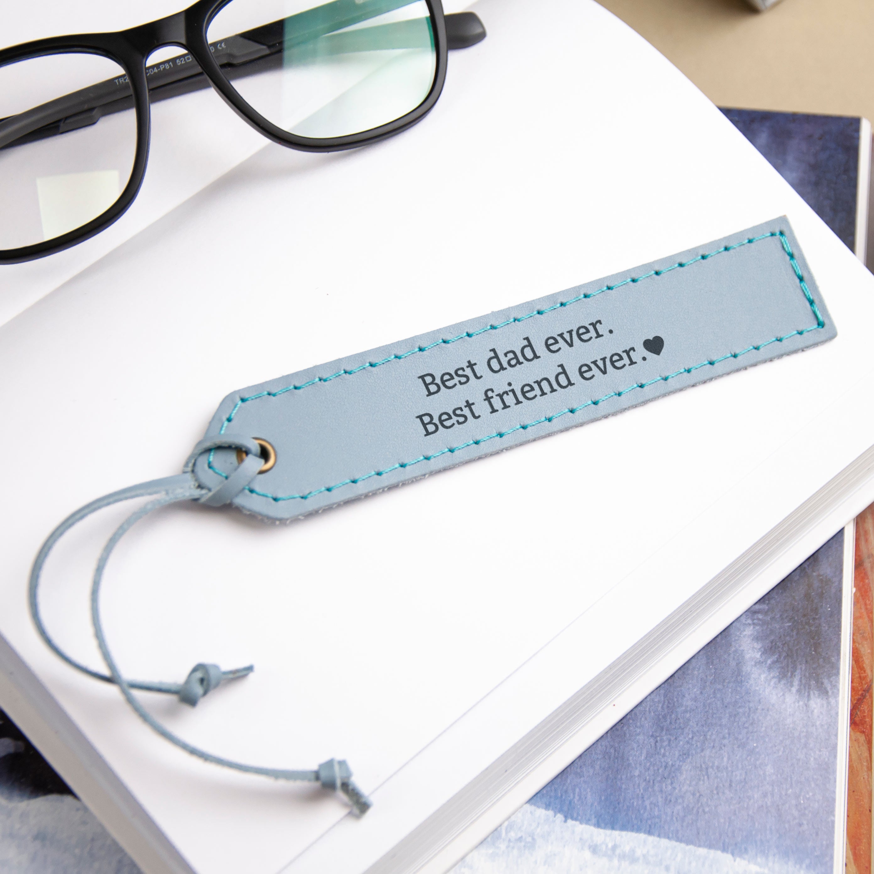 A light blue bookmark with the text "Best dad ever. Best friend ever." is placed on top of a white book.