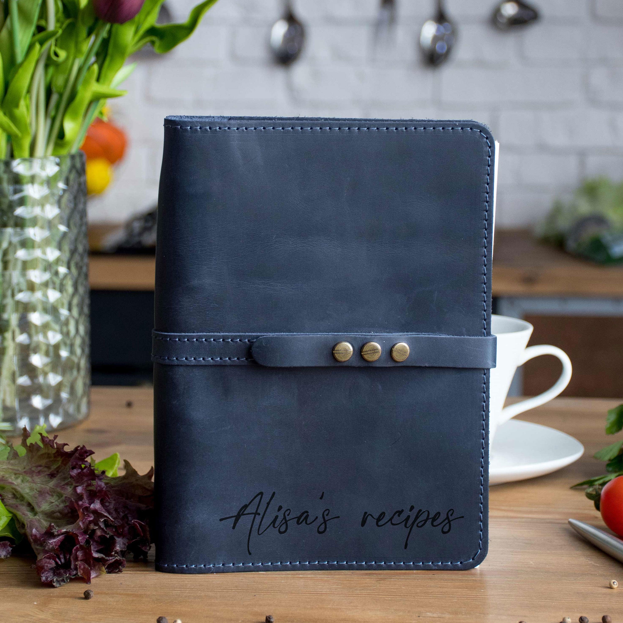 A black leather recipe book with the name "Alison's recipes" printed on it, placed on a wooden table next to a vase of flowers and a cup.