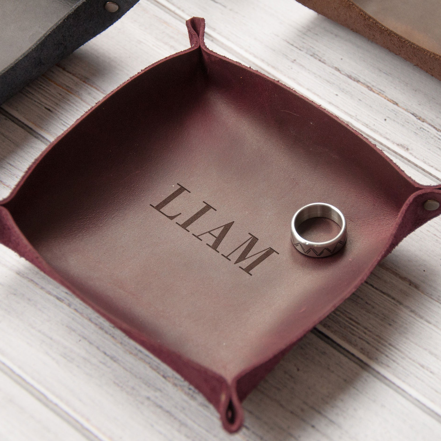 A maroon leather tray with the name "Liam" engraved on it, along with a silver ring placed on top of it.