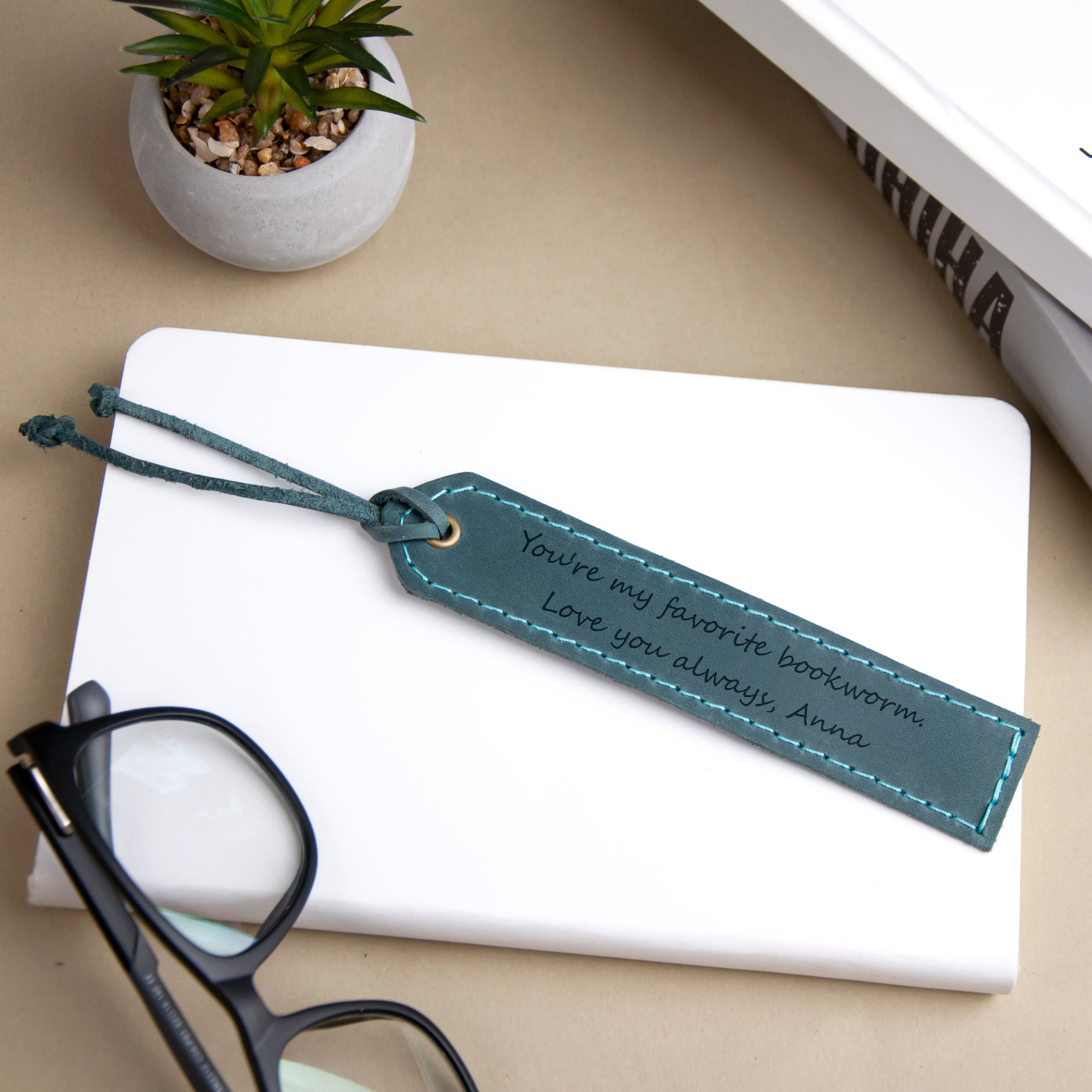 A white notebook, a pair of glasses, and a small potted plant are arranged on a desk, with a personalized bookmark featuring a message about a favorite book Worm on the notebook.