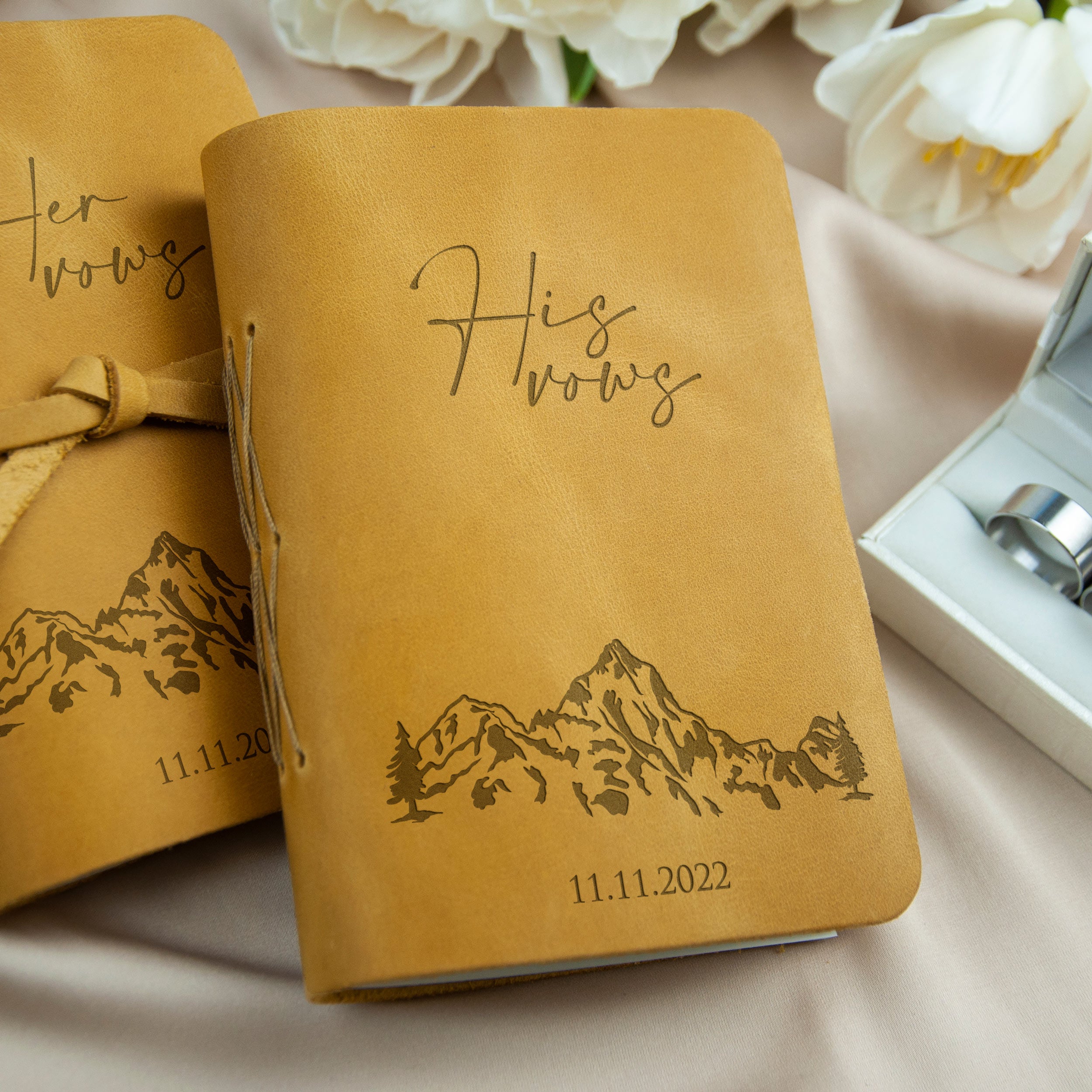 two leather-bound notebooks with a mountain landscape design and the text "His Wows" printed on them. The notebooks are placed on a surface with white flowers in the background.