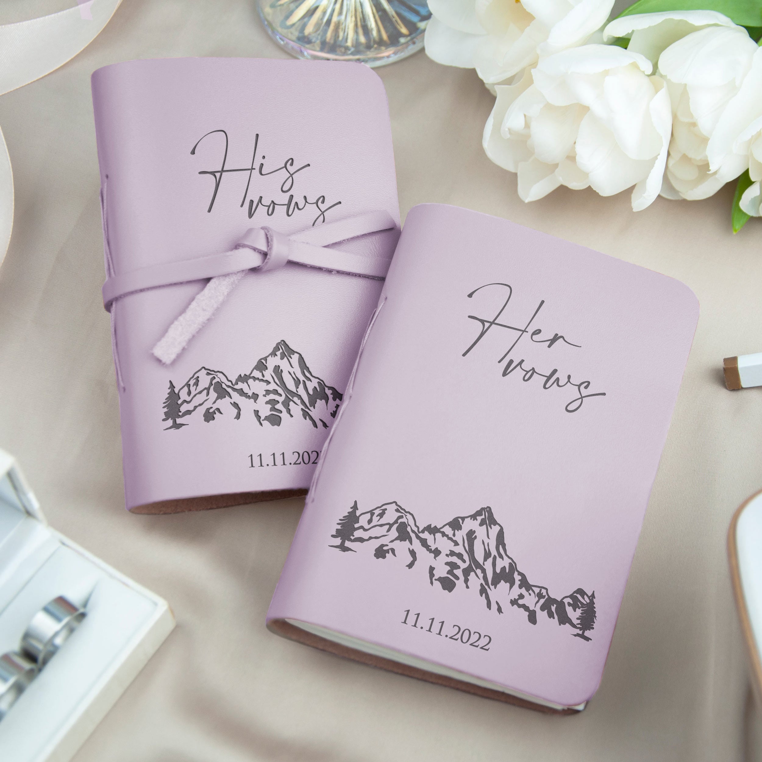 two purple notebooks with the text "Husband" and "Wife" printed on them, along with a mountain illustration. The notebooks are placed on a table with a bouquet of white flowers in the background.