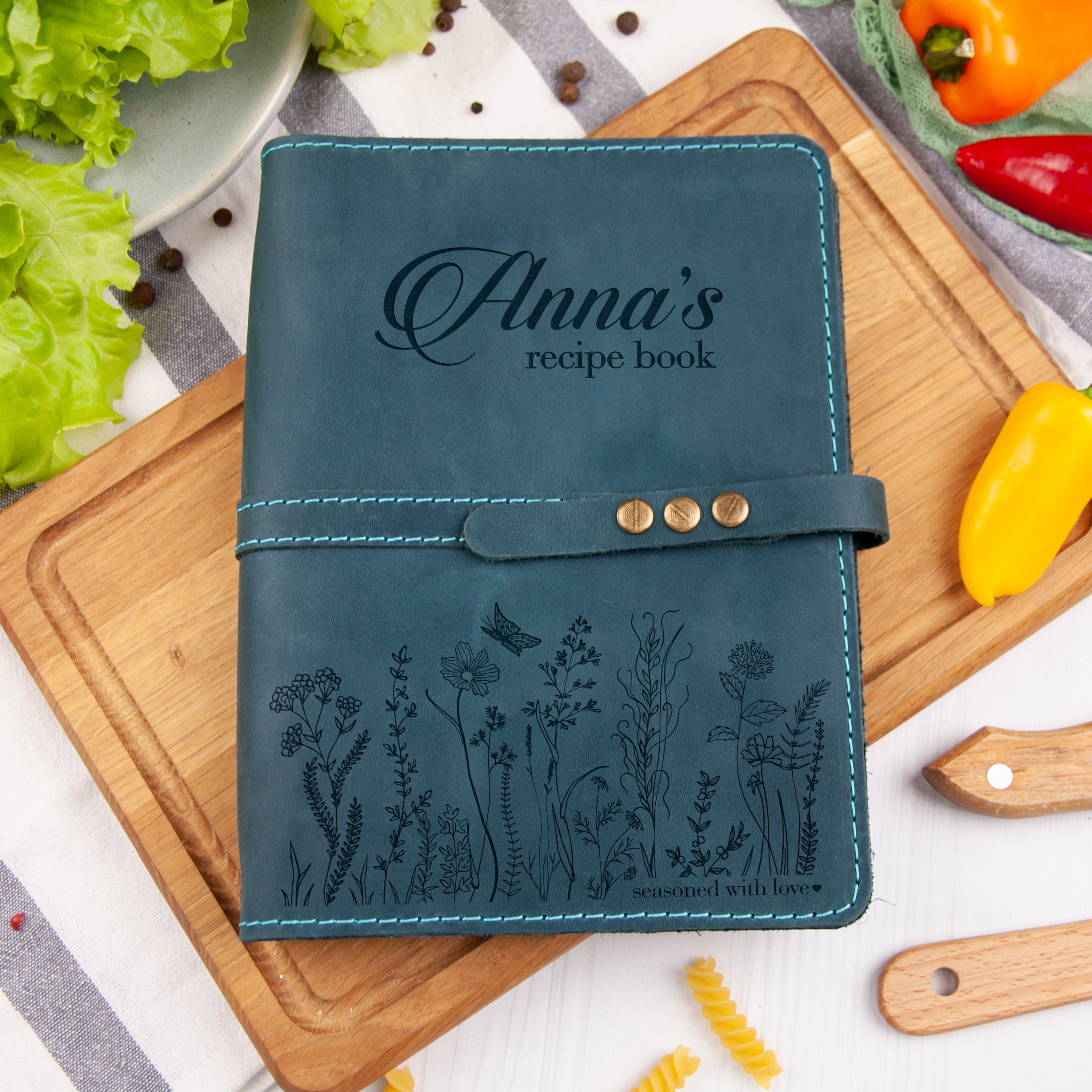 A personalized recipe book with a floral design, placed on a wooden cutting board surrounded by fresh vegetables and a wooden spoon.