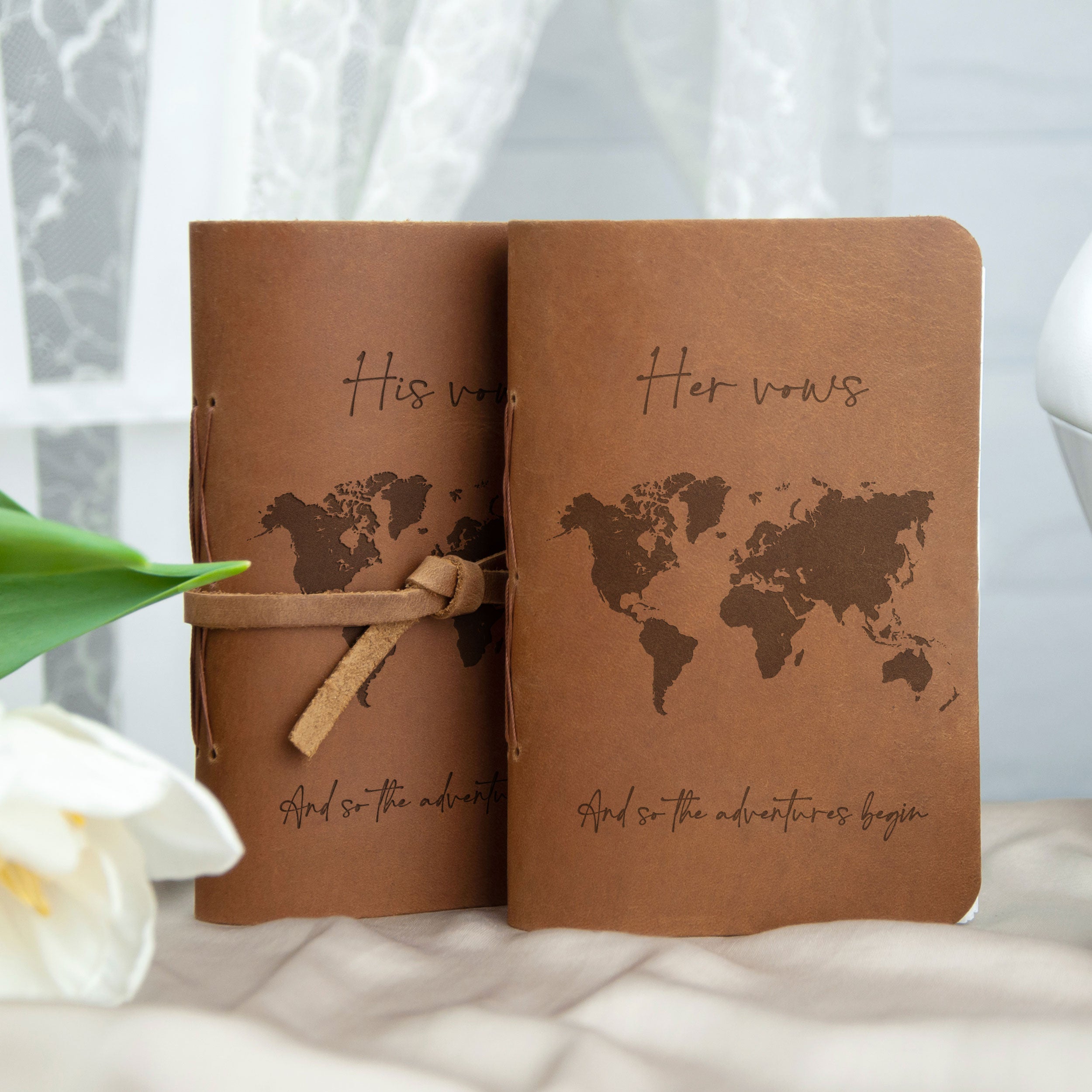 Two brown leather-bound notebooks with a world map design and the text "Her vows" printed on them, placed on a white surface with a green leaf and a white flower in the background.