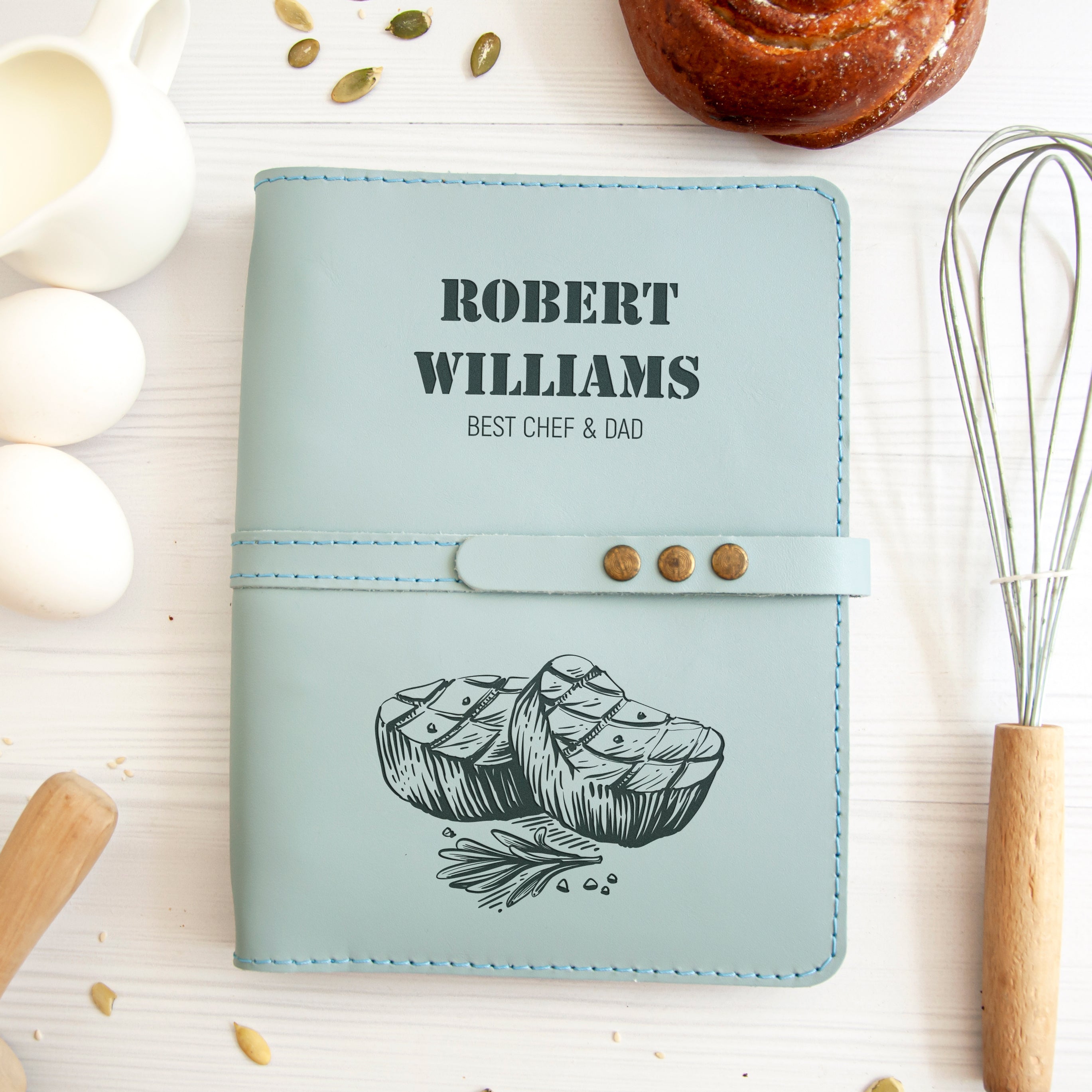 A light blue leather-bound book titled "Robert Williams Best Chef & Dad" with a graphic of a loaf of bread and a chef's hat on the cover, along with a wooden spoon and a white ceramic pitcher.