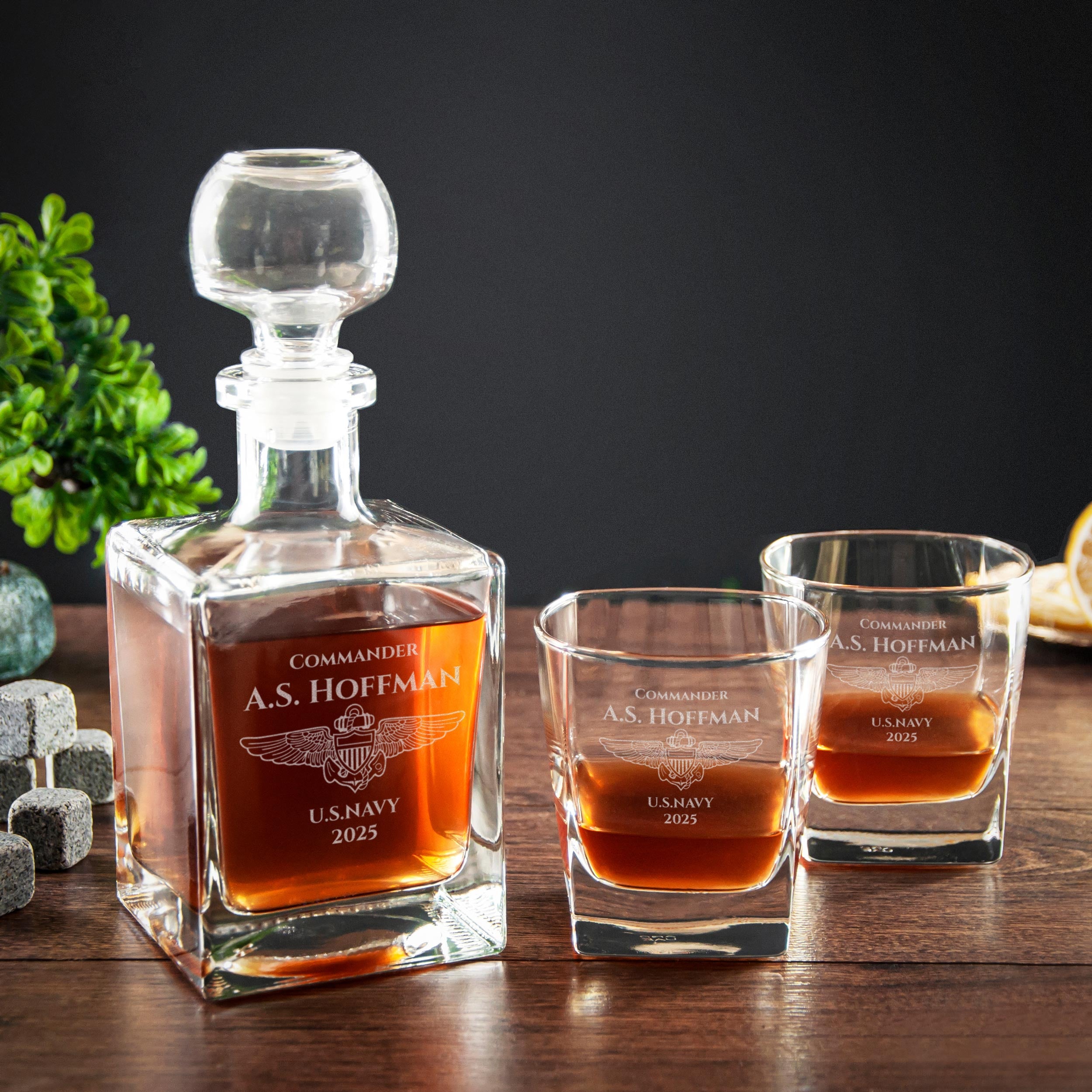 A glass decanter with a bottle of whiskey and two glasses on a wooden surface, with a plant in the background.