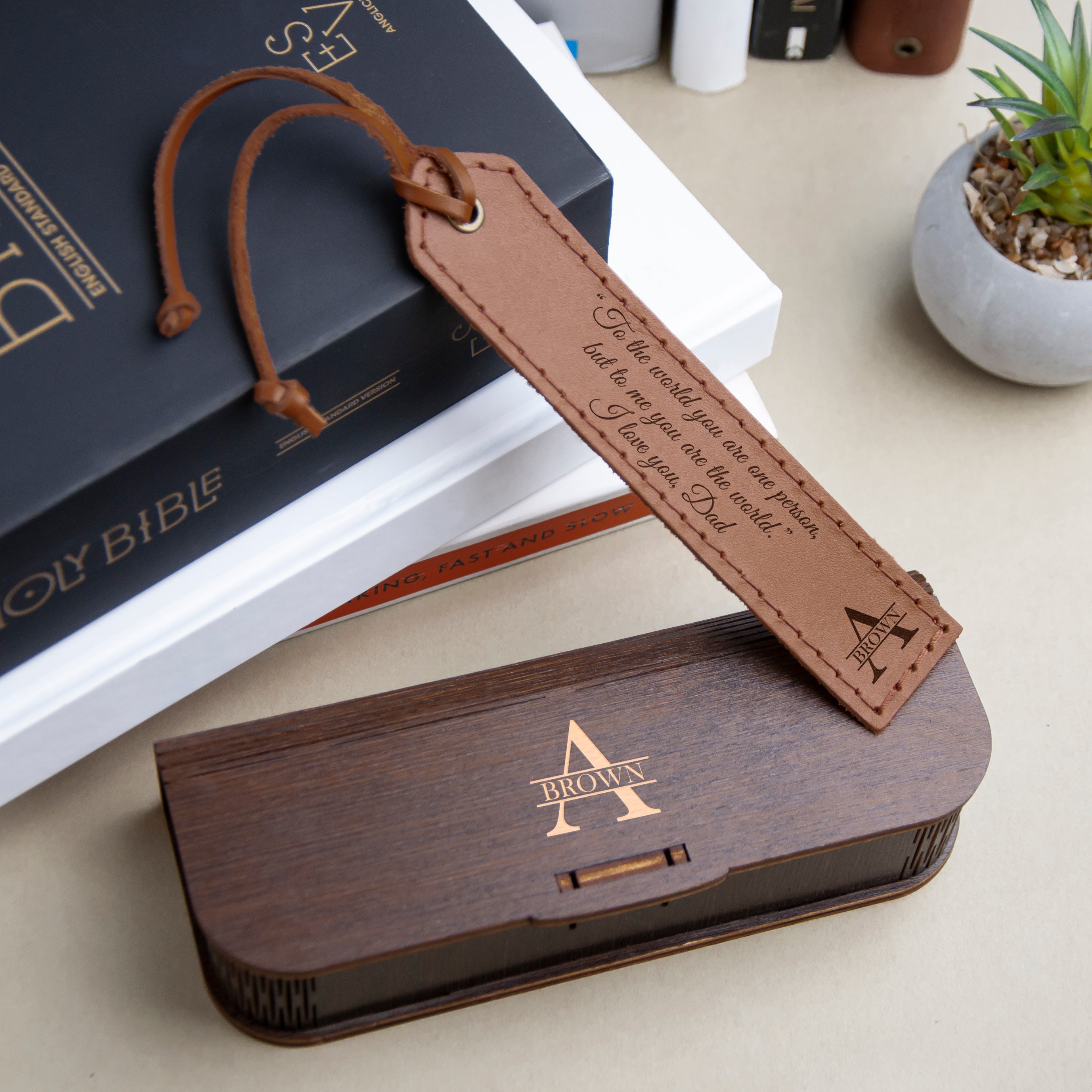 a leather bookmark with a personalized message, a wooden box with a personalized name, and a stack of books on a table.