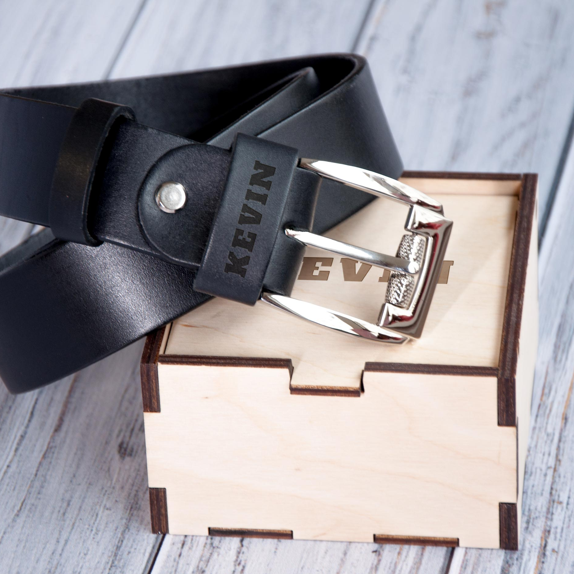 A black leather belt with a silver buckle is placed on top of a wooden box with the name "KEVIN" printed on it.