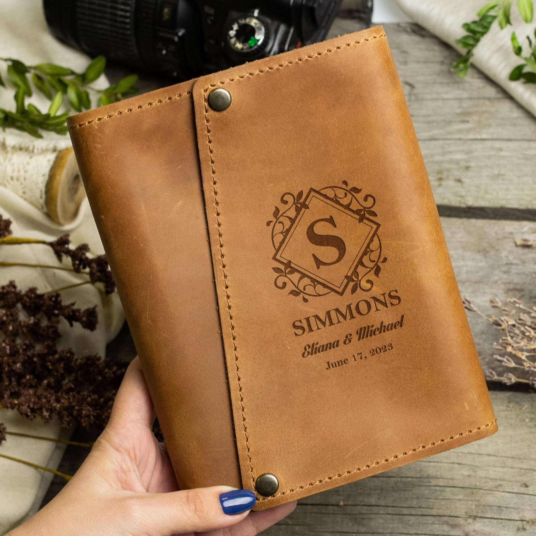 A person's hand holding a brown leather-bound book with a monogrammed "S" and text that reads "SIMMONS Eliana & Michael June 17, 2013".