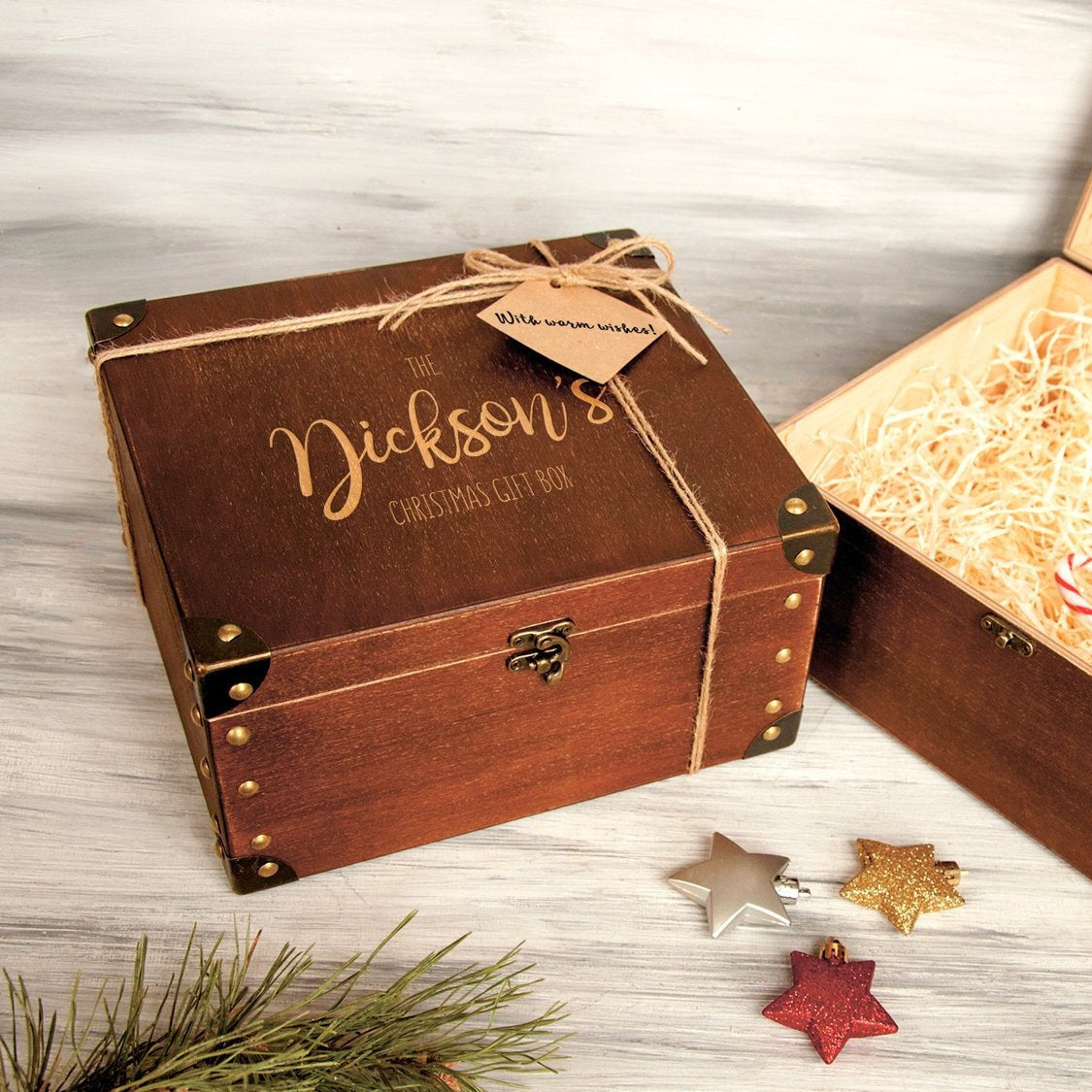 A wooden gift box with a label that says "The Jackson's Christmas Gift Box" and a tag that says "with a wish!" is placed on a wooden surface, accompanied by some ornaments and pine branches.