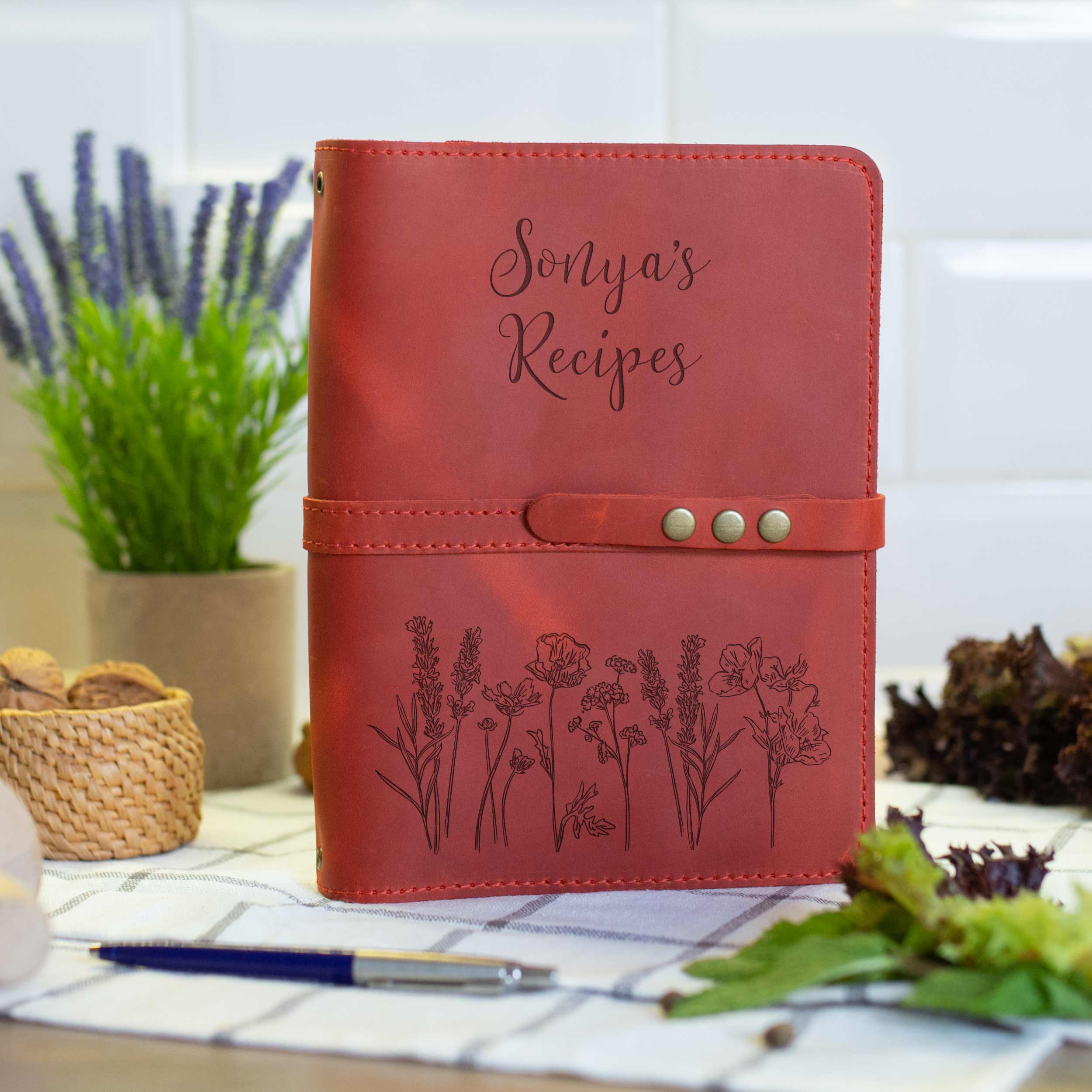 A red leather recipe book with the title "Sonya's Recipes" printed on the cover, placed on a table with a potted plant and some flowers in the background.