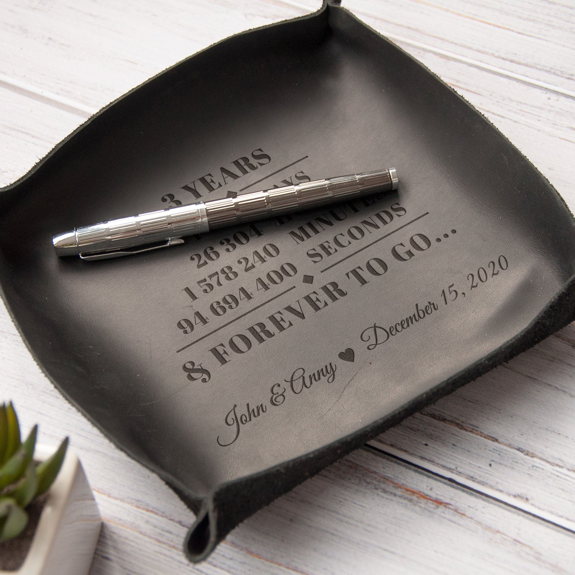 A black leather tray with a personalized message engraved on it, placed on a white surface.