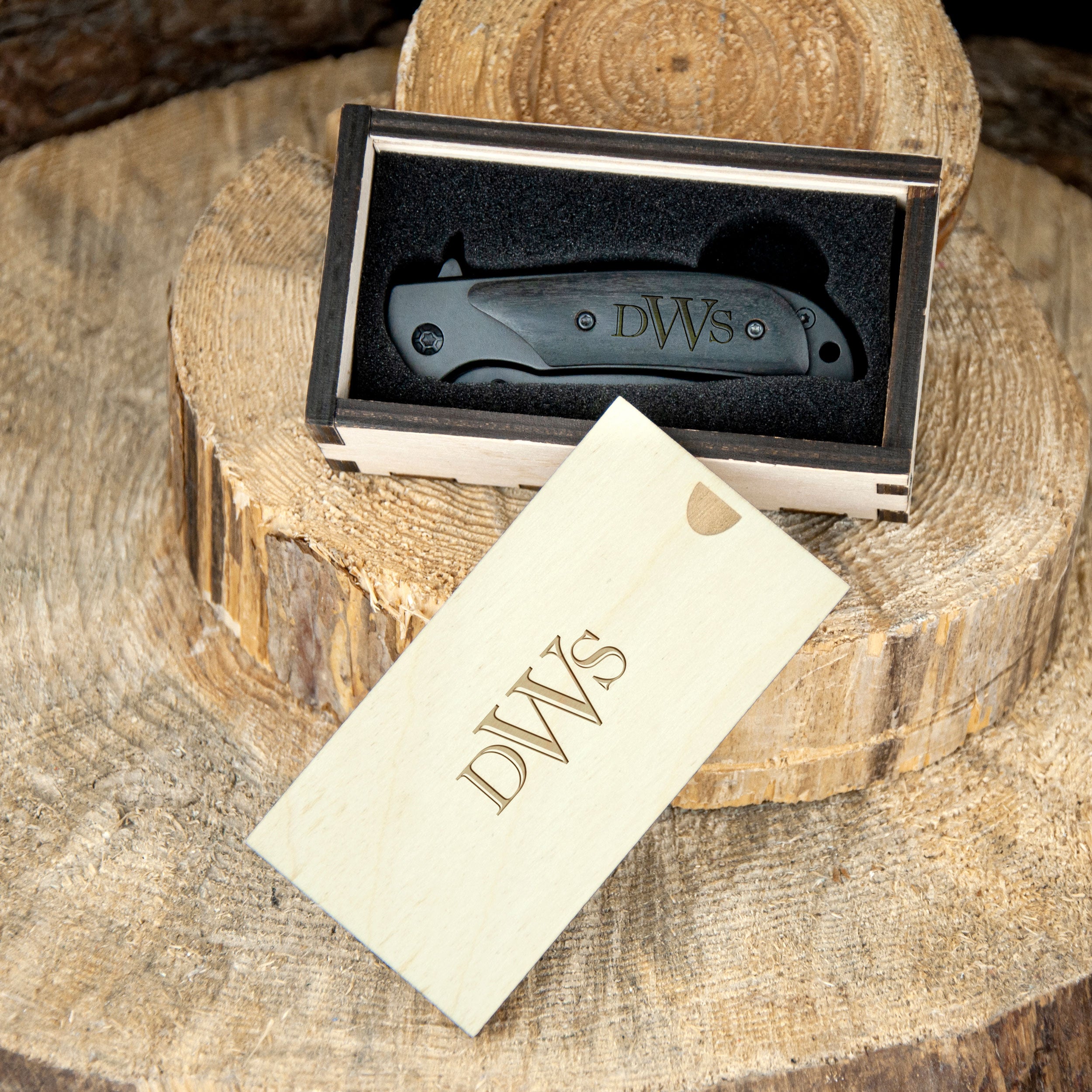 A black folding knife is placed inside a wooden box, with a wooden tag featuring the initials "DWS" on it.