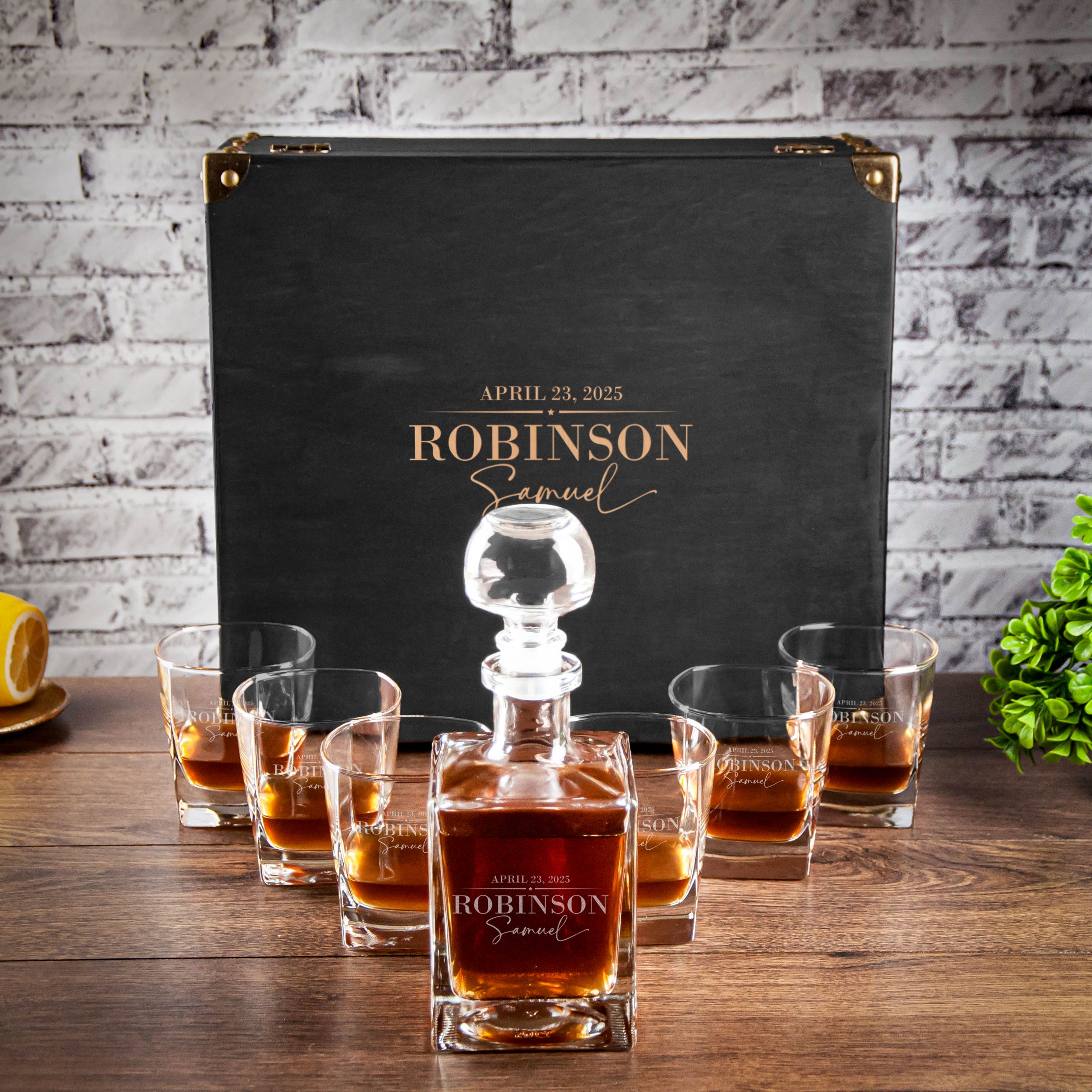 A set of six whiskey glasses with a bottle of whiskey in the center, placed on a wooden table in front of a black briefcase with gold lettering.