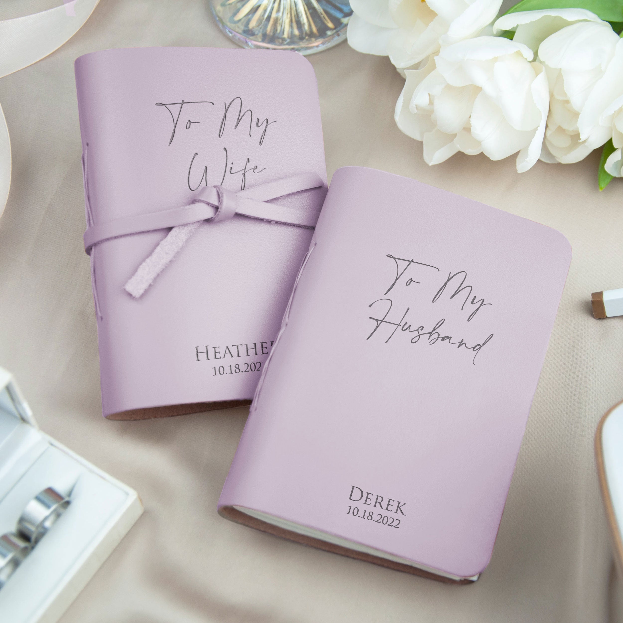 two personalized purple notebooks with the names "To My Wife" and "To My Husband" printed on them, along with the date "10.18.2022". The notebooks are placed on a table with a vase of white flowers in the background.