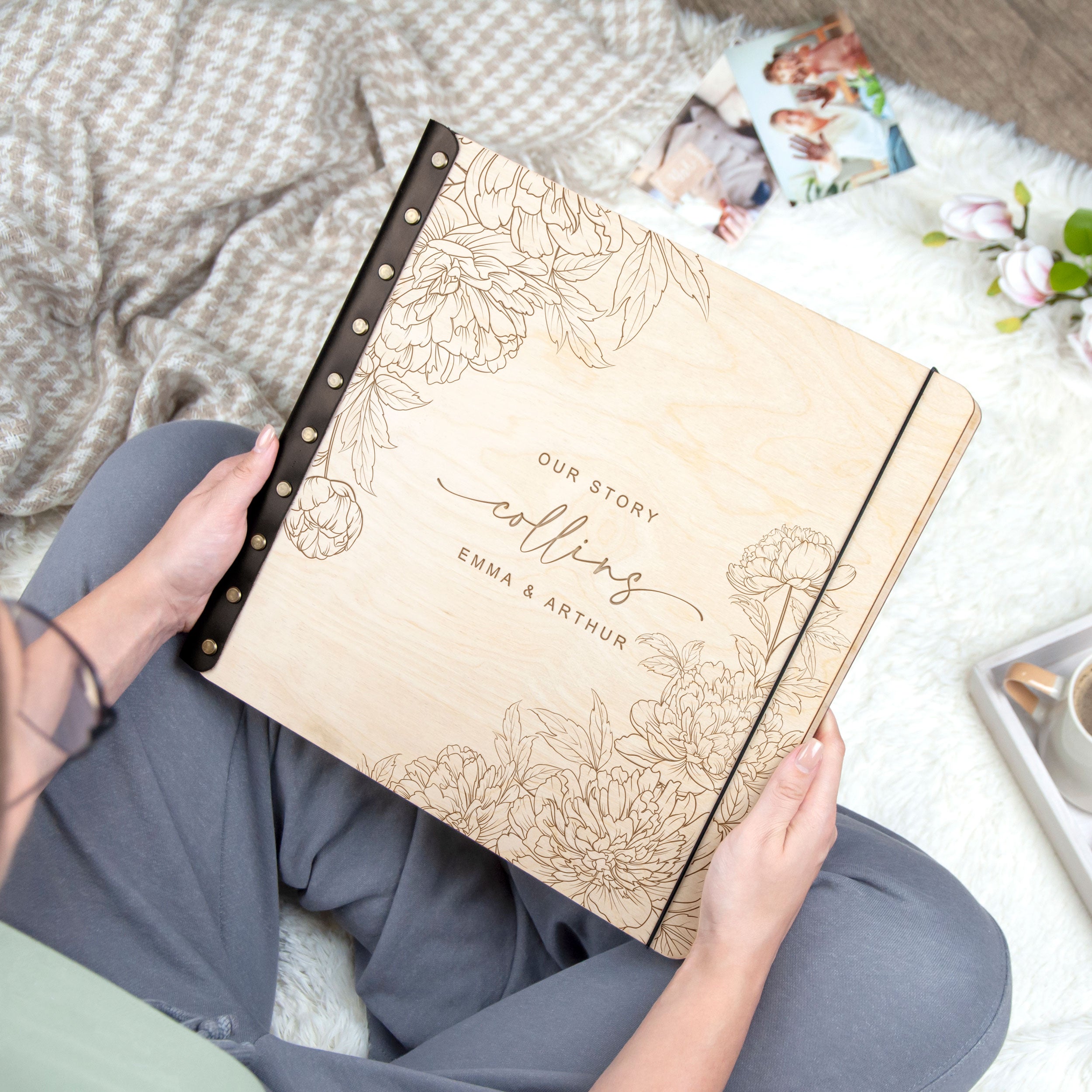A person is holding a wooden photo album with a floral design, and there are some flowers and a cup visible in the background.