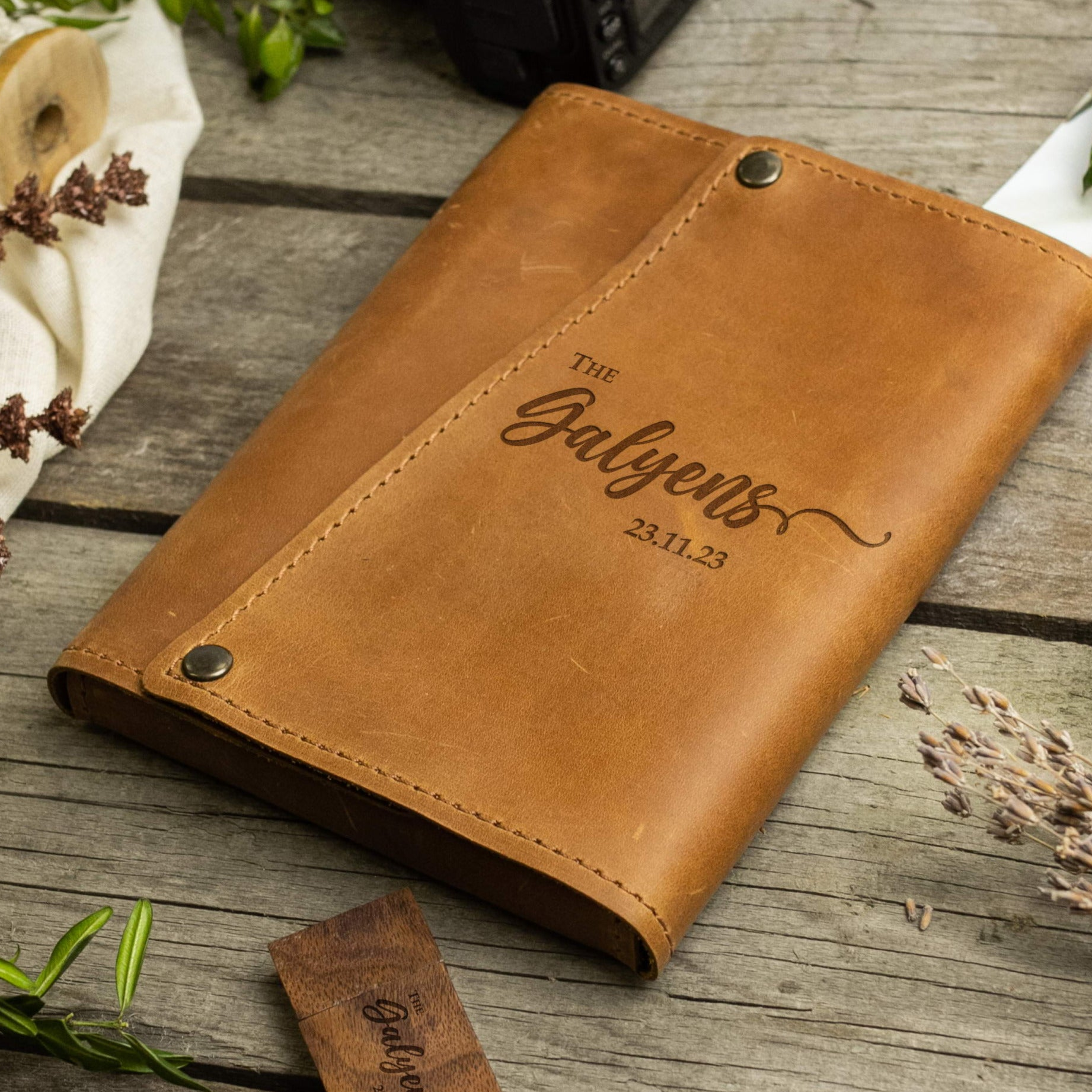 A brown leather-bound notebook with the name "The Balys" printed on it, sitting on a wooden surface with some plants and other items in the background.