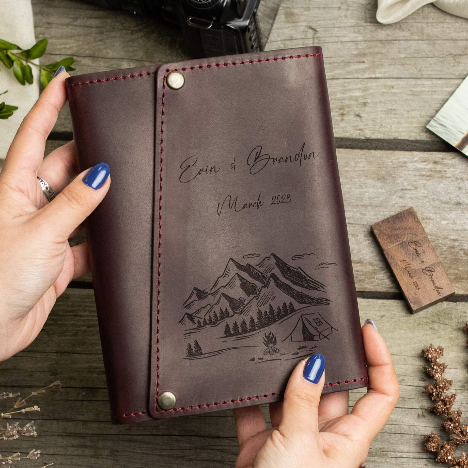 A person's hands holding a brown leather-bound notebook with a mountain landscape and camping scene printed on the cover.