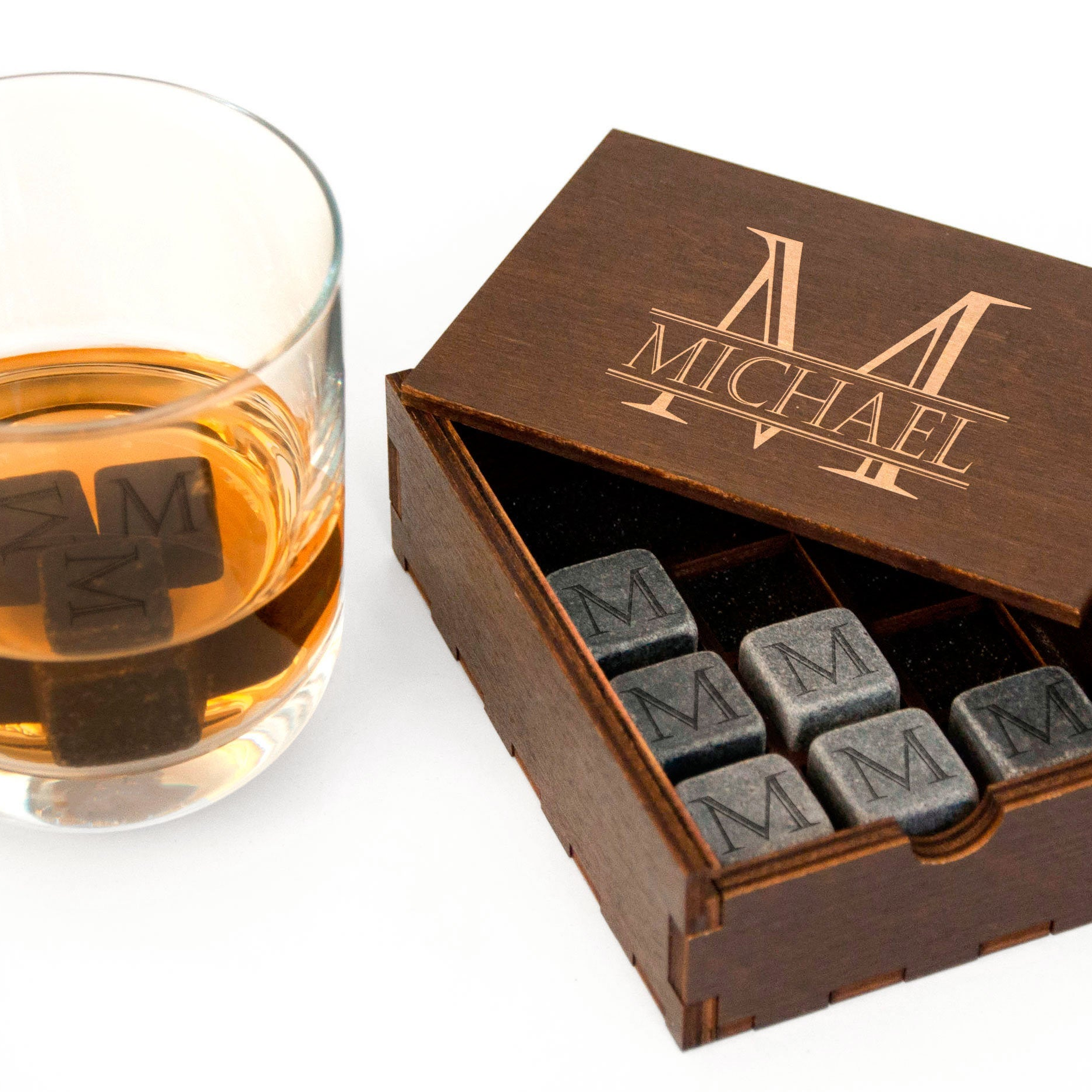 a wooden box with a glass of amber liquid and a set of gray stone cubes with the initials "M" on them.