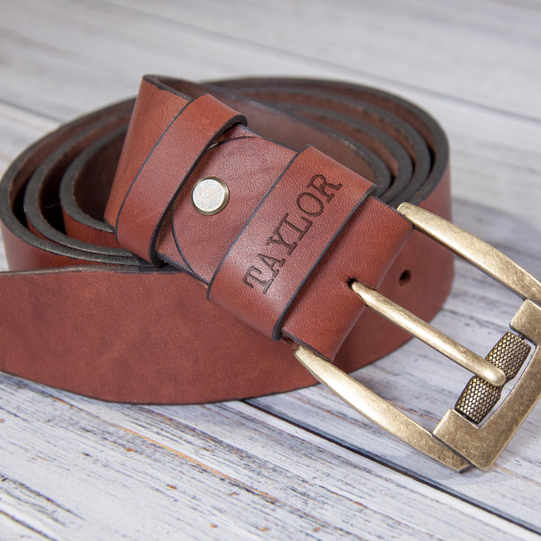 A brown leather belt with a personalized name engraved on it, resting on a wooden surface.