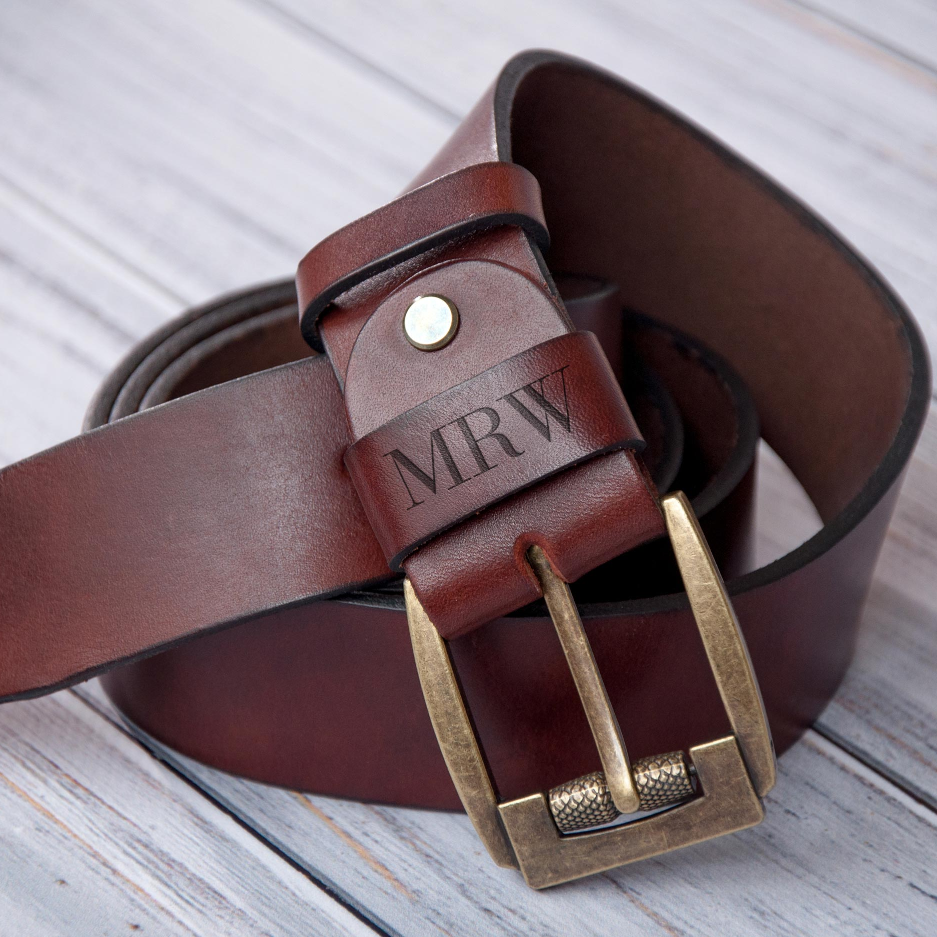 A brown leather belt with a personalized monogrammed buckle, resting on a wooden surface.