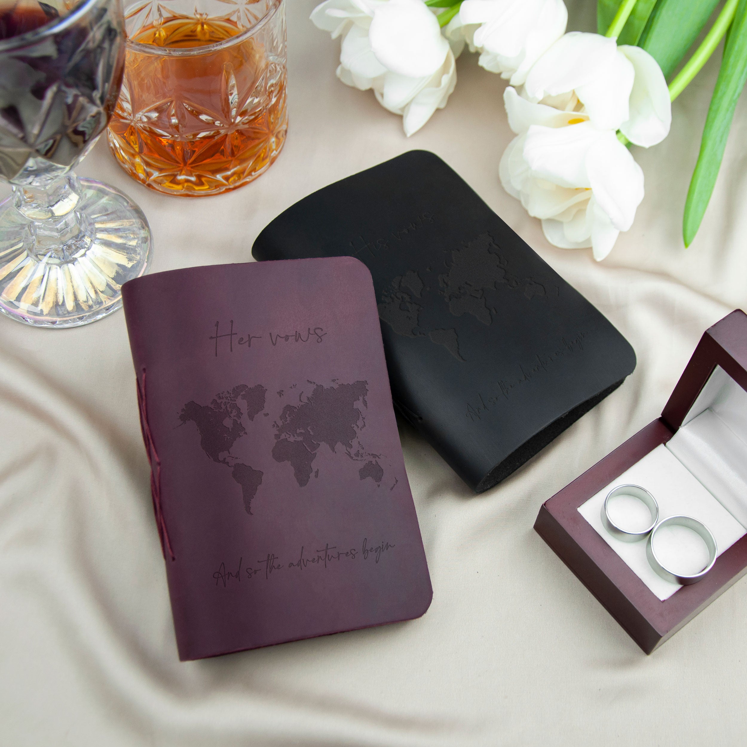 a purple leather-bound book with a world map design, a glass of amber liquid, a bouquet of white flowers, and a small wooden box containing a pair of silver wedding rings.