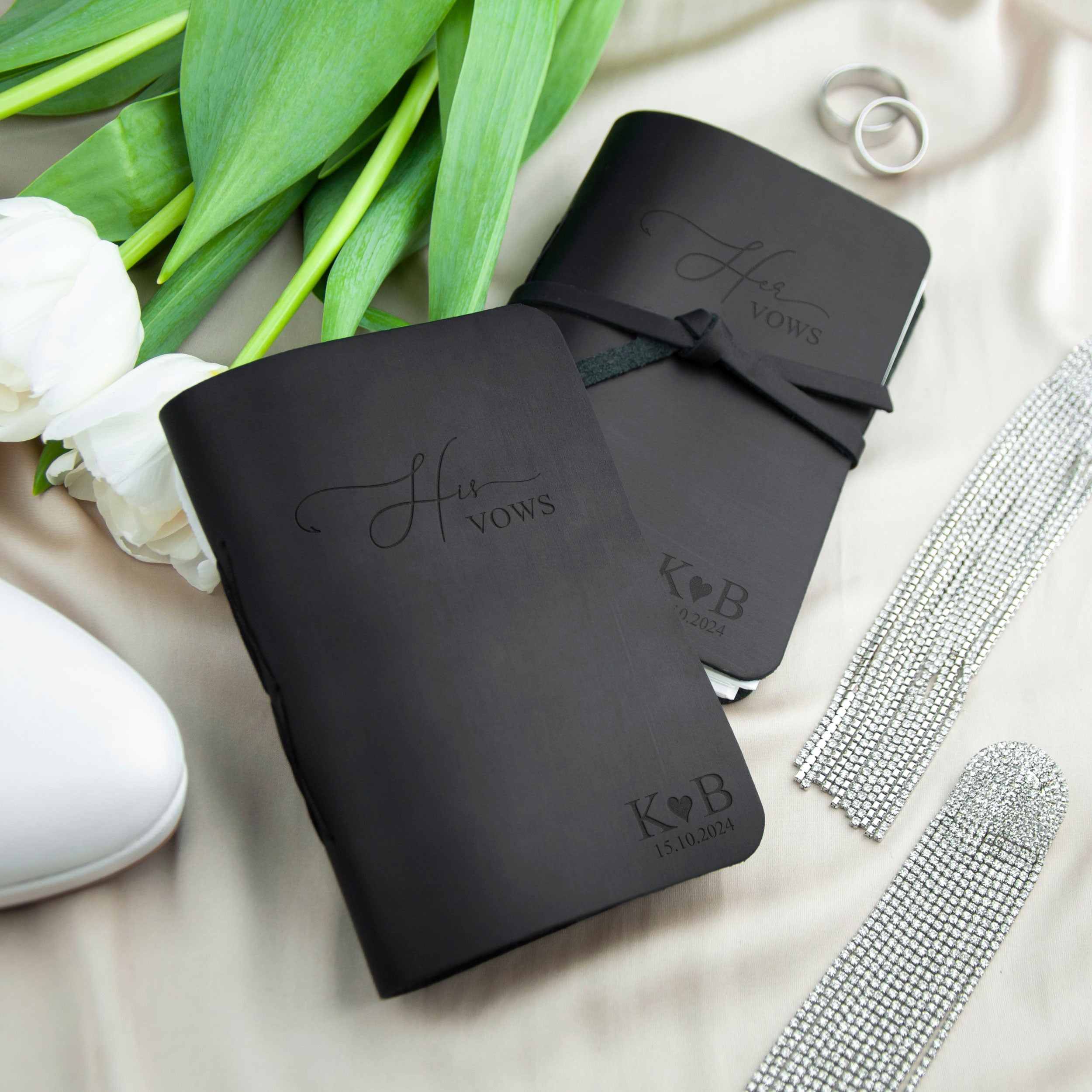 a black leather-bound book with the text "His Vows" printed on it, along with a silver ring and a silver necklace. The book is placed on a white surface, and there are some green leaves and a white flower nearby.