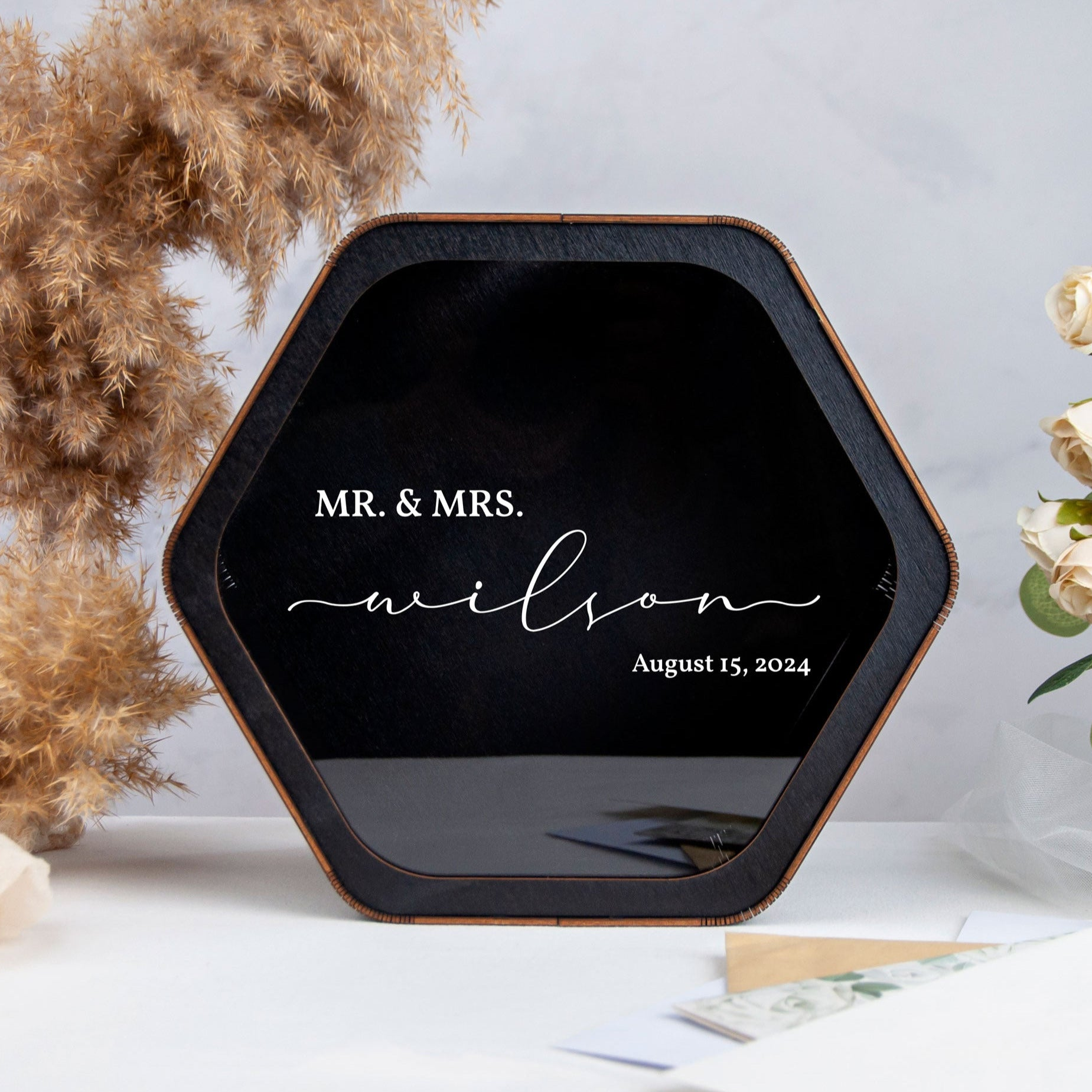 A black hexagonal photo frame with the names 'Mr. & Mrs. Wilson' and the date 'August 15, 2024' printed on it, placed on a white surface next to a potted plant.